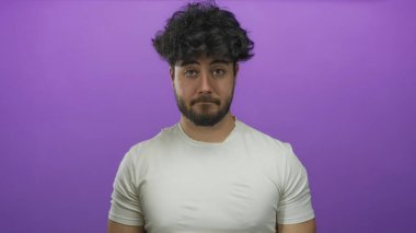 Beyaz tişörtlü, sakallı, genç İspanyol adam canlı mor arka plana karşı duruyor. Düşünceli bir yüz ifadesi var. Modern gündelik stil ve izolasyon kavramı sergiliyor.