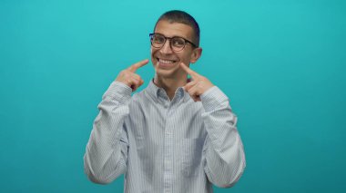 Gözlüklü ve çizgili gömlekli bir adam mavi arka planda gülüşünü gösteriyor, izole edilmiş bir stüdyoda güven ve neşe yayıyor..
