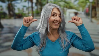 Mavi kazaklı son sınıf öğrencisi kadın, kendine güveni somutlaşmış, kendine güveni somutlaşmış ve neşeli bir açık hava ruhu olan palmiye ağaçlarıyla dolu canlı bir sokakta kendini işaret ediyor..