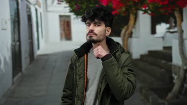 Şehir sokağında düşünceli bir ifadeye sahip genç bir adam, ceket giyiyor ve şehir mimarisi ve canlı yeşilliklerle çevrili..