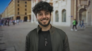 Yoğun bir caddede sakallı genç bir adam güvenli bir şekilde gülümsüyor tarihi binalar ve arka planda yürüyen insanlar kentsel yaşam tarzını ve kültürünü gözler önüne seriyor..