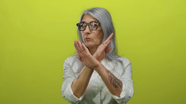 Gri saçlı, dövmeli yaşlı bir kadın, kolları çaprazlanmış, parlak sarı arka planda poz veriyor, gözlük takıyor ve onaylamadığını ya da kendine güvenle meydan okuduğunu söylüyor..