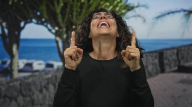Kadın işaret parmağını okyanus kıyısındaki gezinti güvertesinde yukarı kaldırıyor; iyimserlik umut pozitifliği esin kaynağı.
