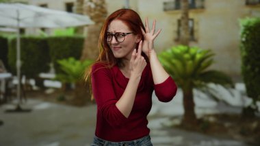 Şehir sokaklarında kızıl saçlı ve gözlüklü bir kadın palmiye ağaçlarıyla çevrili büyük bir şemsiye altında dinleme hareketi yapıyor..