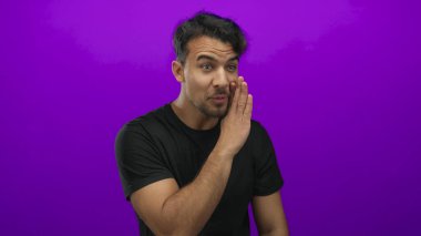 Genç İspanyol bir adam, izole edilmiş mor arka planda fısıldıyor. Siyah gömlekli, neşeli bir ifadeyle neşe ve gizlilik gösteriyor.