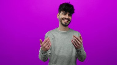 Genç İspanyol bir adam gülümsüyor ve elleri ile işaret ediyor. İzole edilmiş, pembe bir arka planı var. Gri bir süveter giyiyor.