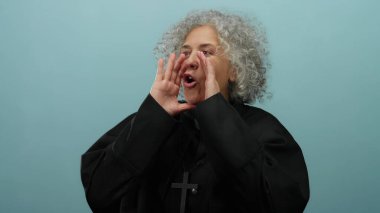 Dini cüppeli, kıvırcık gri saçlı yaşlı bir kadın, dışa kapalı mavi arka planda, anlamlı jestler ve yüz ifadesi yoluyla inancını ve duygularını ifade eden tutkulu bir şekilde konuşuyor.