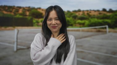 Tenis kortunda gülümseyen, bulanık doğal arka planda süveter giyen, gençlik neşesi ve dinginliği yakalayan bir kadın..