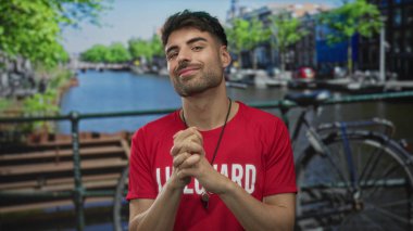 Kanal kenarındaki bisiklet parkı ve nehir kenarındaki binalarla kırmızı cankurtaran gömleği giyen adam ellerini göğsüne yaklaştırıyor..