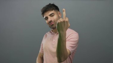 Pembe polo tişörtlü genç İspanyol adam gri stüdyoda kameraya bakan orta parmağını gösteriyor..