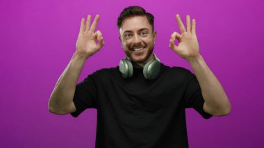 Kulaklıklı genç adam gülümsüyor ve parlak pembe bir arka plana el hareketi yapıyor. Kendine güveni ve pozitifliği var..