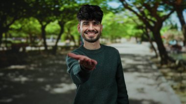 Güneşli bir günde, gölgeli ağaçların altında dostane ve davetkar bir atmosfer yaratan, yemyeşil bir park ortamında gülümseyen ve elini uzatan İspanyol bir adam..