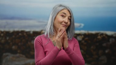 Gri saçlı yaşlı bir kadın açık deniz kenarındaki gezinti alanında pembe bir gömlek giyerek sahilde ve okyanus manzarasında dövmelerini sergiliyor..