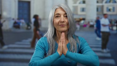 Gri saçlı yaşlı bir kadın hareketli bir açık sokakta duruyor, mavi giyinmiş ve hareketli bir şehir ortamında bulanık yayaların arasında kendini ifade ediyor..