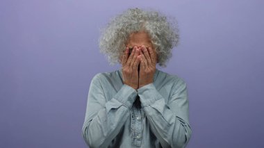 İzole edilmiş mor arka plana karşı duygusal jestlerle yüzünü kaplayan gri saçlı yaşlı bir kadın..