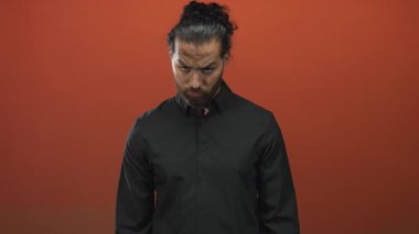 Siyah gömlek giyen ve kapalı gözlü bir adam turuncu bir stüdyo çekiminde aşağı bakıyor; içe dönük sakin.