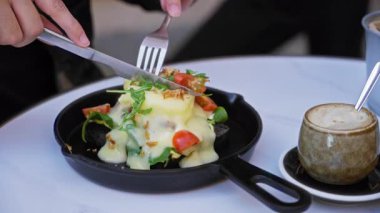 Güneşli bir kafeteryada kapuçinolu bir masada yumurtalı Benedikt yiyen kişi canlı salata ve kremalı sos sergiliyor..