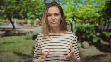 Genç beyaz kadın, güneş ışığı altındaki ağaçların altındaki yeşil parkta gözleri kapalı el kaldırıyor..