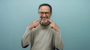 Olgun bir adam mavi bir arka plan karşısında durup gülümsemesini işaret ederek, jesti ve yüz ifadesiyle mutluluğu ifade eder..
