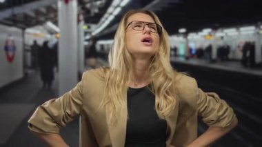 Sarışın ve gözlüklü bir kadın, hareketli bir tren istasyonunda karnını sıkıyor. Parlak iç aydınlatma altında rahatsızlık içinde görünüyor..