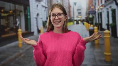 Pembe süveterli gözlüklü genç sarışın kadın şehrin kaldırım taşı sokağında yüzünü gösteriyor. Mutluluk..