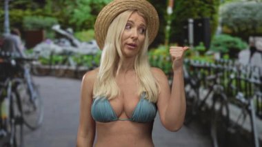 Hasır şapkalı ve mavi bikinili bir kadın başparmağıyla caddede park halindeki bisikletlerle yan yana duruyor..