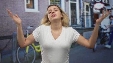 Kadın çıplak kollarıyla omuz silkiyor ve sokakta park halindeki bisiklet ve tuğla binanın yanında avuç içi açık duruyor..
