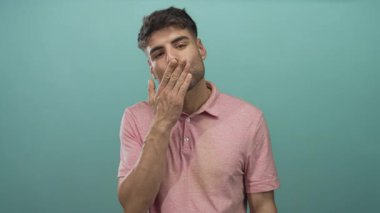 Pembe polo tişörtlü bir adam, turkuaz bir stüdyo ortamında nazikçe elini uzatarak öpücük savurur..