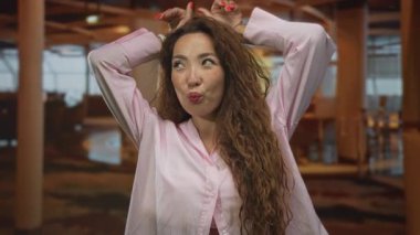 Elleri başının üstünde, neşeli bir kulak hareketi yapan ve yapıda dudaklarını büzen, oyuncu bir doğallık gösteren bir kadın..