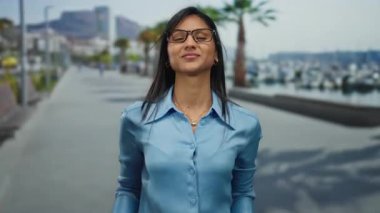 Gözlüklü genç Latin kadın, güneşli deniz kıyısında, arka planda palmiye ağaçları ve plajları olan neşeli bir jest yapıyor..