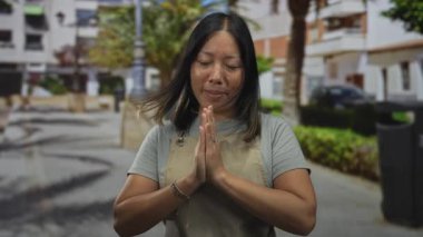 Palmiye ağaçları ve konut binasının yanındaki bir sokakta dua jestinde elleri birbirine bastırılmış genç İspanyol kadın, sakin minnet yansıması..