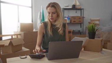 Yeni evinde dizüstü bilgisayar üzerinde çalışırken hesap makinesi kullanan sarı saçlı genç bir kadın. İç dekorasyonu ve saksı bitkisi olan oturma odasındaki kutuları taşıyor..