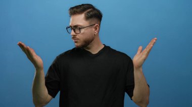 Sakallı ve gözlüklü genç adam mavi bir arka planda, kararsızlık tasvir eden ellerine bakıyor ve.