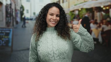 Dışarıdaki terasta kıvırcık saçlı, süveter giyen, kafelerle çevrili sokakta duygularını ifade eden ve gündüzleri arka planda olan kadınlar..