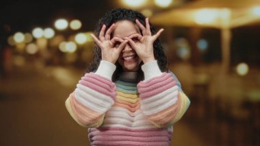 Elleriyle gözlük hareketi yapan bir kadın aydınlık bir sokakta renkli bir süveter içinde neşeli bir şekilde gülümseyen açık hava havası veren bir kadın..