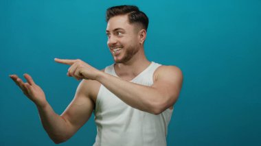 Beyaz atletli sakallı genç adam gülümsüyor ve açık bir eli işaret ediyor. Mavi arka plana karşı. Pozitif ve davetkar bir sunum yapıyor..