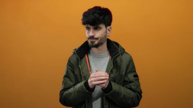 Yeşil ceketli genç İspanyol bir adam izole edilmiş turuncu bir arka planda dikiliyor. Yüzünde düşünceli bir ifadeyle serin bir ifade ve sımsıkı tutulmuş eller soğuk havayı işaret ediyor..