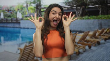 Turuncu tişörtlü gülümseyen İspanyol kadın havuz başında iki eliyle ahşap güvertede 