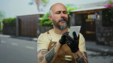 Sakallı bir adam, şehir sokaklarında defter tutuyor, eldiven ve önlük takıyor, motosikletli bulanık bir arka planı var..