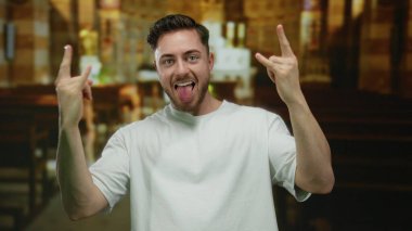 Sakallı genç adam kilisenin içinde taş jestler yapıyor. Beyaz gömlek giyiyor ve ev ortamında heyecan ve ifade gösteriyor..