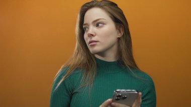 Düşünceli duruş ve tarafsız ifadeyle turuncu stüdyoda akıllı telefonu tutarken kadın yana bakıyor; merak..