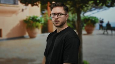 Şehir sokaklarında sakallı genç bir adam güneşli bir günde arka planda bitkilerle şehir yaşam tarzını gözler önüne seriyor..