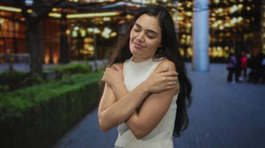 Gece yarısı apartman ışıkları altında çıplak kollarıyla sokakta çıplak kollarını saran bir kadın, kendinden memnun..