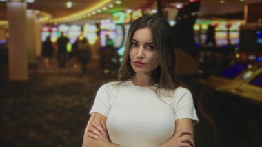 Genç İspanyol kadın kollarını meşgul bir kumarhane binasında kumar makinesinin yanında kavuşturuyor..