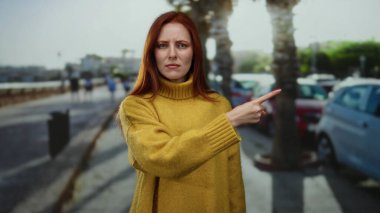 Şehir caddesinde kendine güvenen bir ifadeyle park etmiş arabalarla ve güneşli bir gökyüzünün altında palmiye ağaçlarıyla çevrili sarı bir süveter giyen bir kadın..