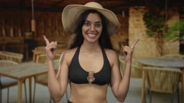 Hasır şapkalı kadın dışarıda teras inşa ederken gülümsüyor ve parmağıyla objeyi işaret ediyor; heyecan.