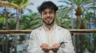 Sakallı ve gözlüklü genç adam güneşli bir parkta gülümsüyor beyaz bir ceket giyiyor, yemyeşil palmiye ağaçlarıyla çevrili, açık elleri ve sakin tavırları var..