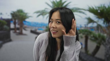 Palmiye ağaçlarıyla çevrili ve rahat bir süveter giyen, sakin bir sahil gününün neşeli ruhunu yakalayan, manzaralı bir sahil gezisinde dikkatlice dinleyen bir kadın..