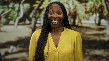 Parkta ışıl ışıl gülümseyen bir kadın. Arka planda ağaçlar olan sarı bir gömlek giyiyor. Güneşli bir günde neşe ve doğa sergiliyor..