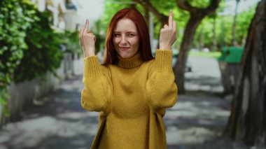 Şehir caddesinde, sarı süveter giyen, ağaçlar ve binalar arka planda sıralanırken kızıl saçlı, hayal kırıklığına uğramış jestler yapan bir kadın..